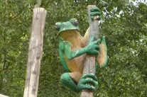 große Froschfigur an einem senkrechten Holzstamm, links daneben noch ein senkrechter Holzstamm, im Hintergrund Baumblätter