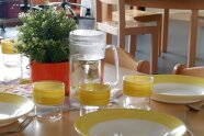 Johanniter gedeckter Tisch mit Tellern, Gläsen und Besteck alles mit gelbem Rand. In der Mitte eine Glaskaraffe und eine kleine Grünpflanze