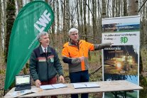 Zwei Herren stehen hinter einem Schild umgeben von Werbetafeln 